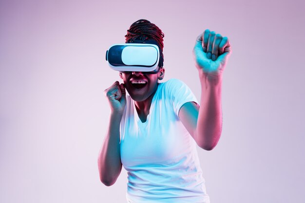 Portret van jonge Afro-Amerikaanse vrouw spelen in VR-bril in neonlicht op verloop achtergrond