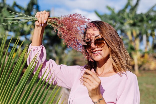 Portret van jonge blanke gelooide vrouw in romantische roze jurk ronde oorbellen zilveren armband en zonnebril met wilde bloemen