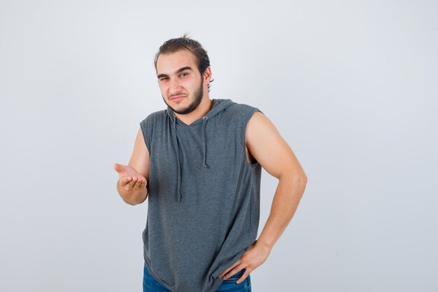 Portret van jonge fit mannelijke spreidende palm op camera in mouwloze hoodie en aarzelend vooraanzicht op zoek