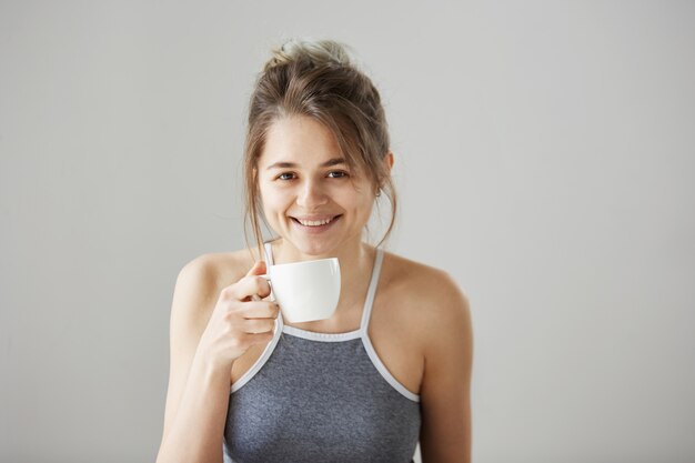 Portret van jonge gelukkig mooie vrouw die lacht bedrijf kopje koffie drinken op ochtend over witte muur.