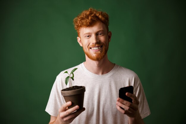 Portret van jonge glimlachende roodharige bebaarde jonge man, met gevlekte plant en mobiele telefoon