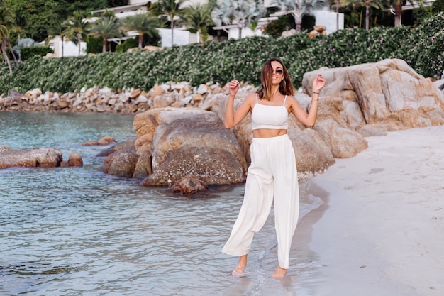 portret van jonge kalme gelukkig Kaukasische fit slanke vrouw in crop cami top en broek alleen op rotsachtig tropisch strand bij zonsondergang