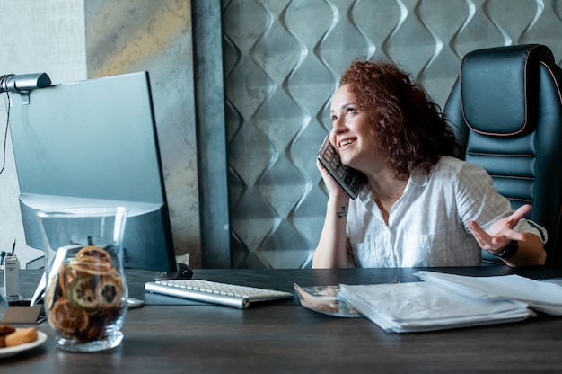 Gratis foto portret van jonge kantoormedewerker vrouw zittend aan een bureau met behulp van rekenmachine als mobiele telefoon plezier glimlachend vrolijk in kantoor