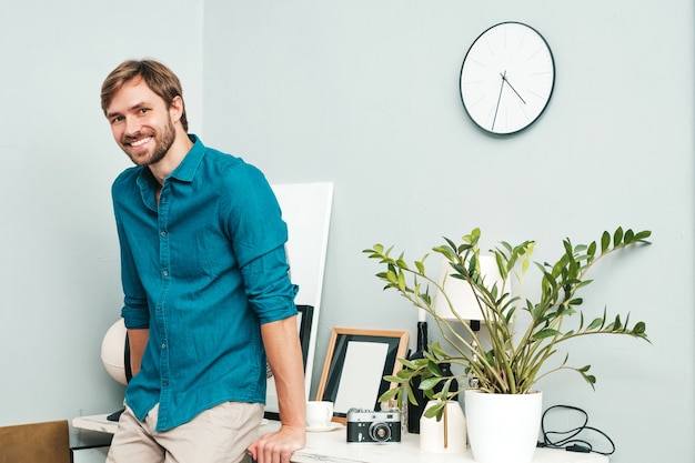 Portret van jonge knappe zakenman. Glimlachend mannetje gekleed in spijkerbroekoverhemd. Bebaard model poseren op kantoor in de buurt van papieren bureau