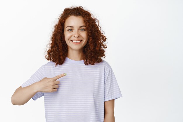 Portret van jonge krullende vrouw met rood haar op witte glimlach, wijzende vinger en zelfverzekerd kijkend, staande in tshirt op wit.