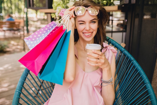 Portret van jonge lachende gelukkig mooie vrouw met opgewonden gezicht expressie zitten in café met boodschappentassen koffie drinken, zomer mode outfit, hipster stijl, roze katoenen jurk, trendy kleding