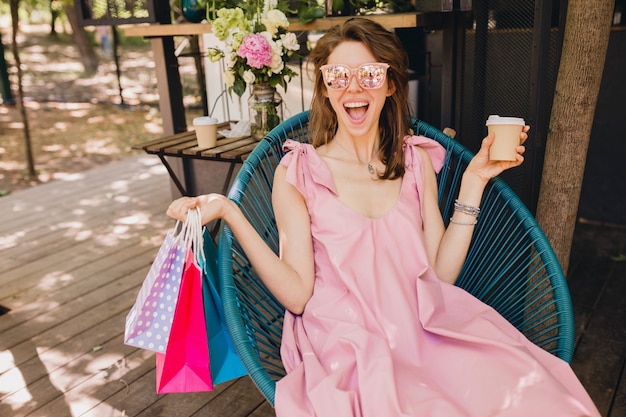 Portret van jonge lachende gelukkig mooie vrouw met opgewonden gezicht expressie zitten in café met boodschappentassen koffie drinken, zomer mode outfit, roze katoenen jurk, trendy kleding