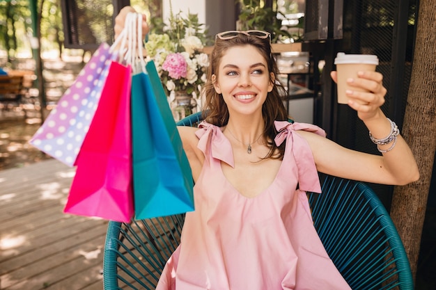 Portret van jonge lachende gelukkig mooie vrouw met opgewonden gezicht expressie zitten in café met boodschappentassen koffie drinken, zomer mode outfit, roze katoenen jurk, trendy kleding