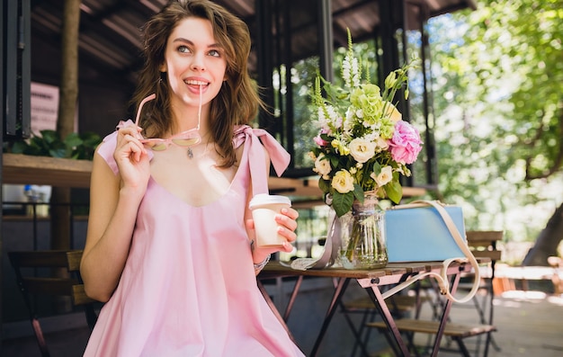 Portret van jonge lachende gelukkig mooie vrouw met zittend in café koffie drinken, zomer mode outfit, hipster stijl, roze katoenen jurk, trendy kledingaccessoires