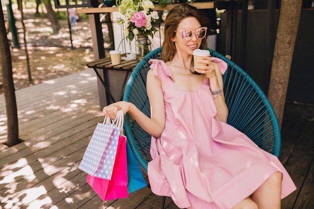 Portret van jonge lachende gelukkig mooie vrouw zitten in café met boodschappentassen drinken koffie, zomer mode outfit, roze katoenen jurk, trendy kleding