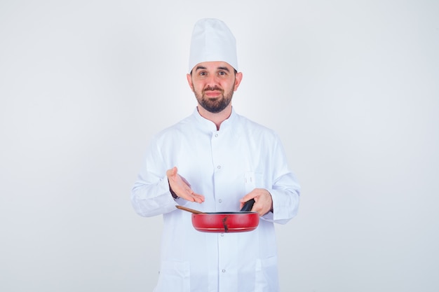 Portret van jonge mannelijke chef-kok die lege koekenpan met houten lepel in wit uniform toont en neergeslagen vooraanzicht kijkt