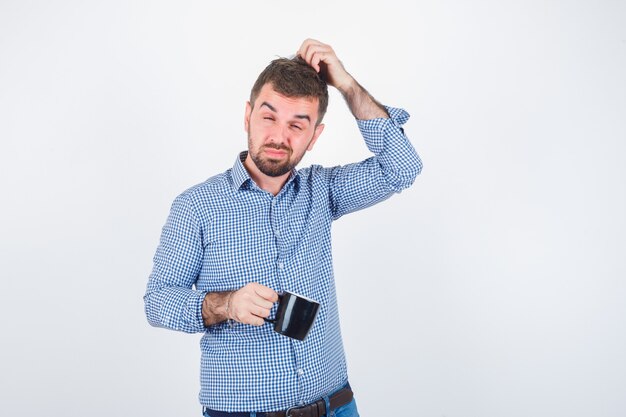 Portret van jonge mannelijke hoofd krabben terwijl cup in shirt, spijkerbroek en peinzende vooraanzicht kijken