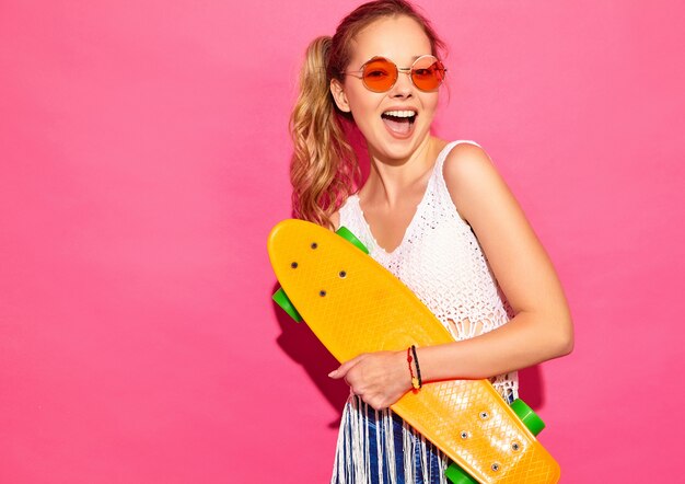 Portret van jonge modieuze glimlachende blonde vrouwen met stuiverskateboard. vrouwen in de zomer witte jurk poseren in de buurt van roze muur