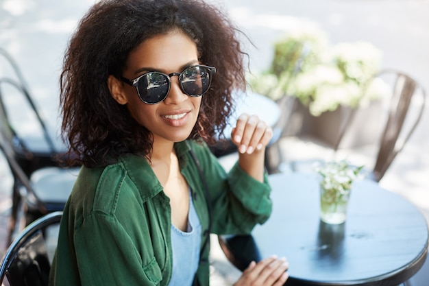 Portret van jonge mooie afrikaanse vrouw in sungasses glimlachend rusten ontspannen in café op terras.