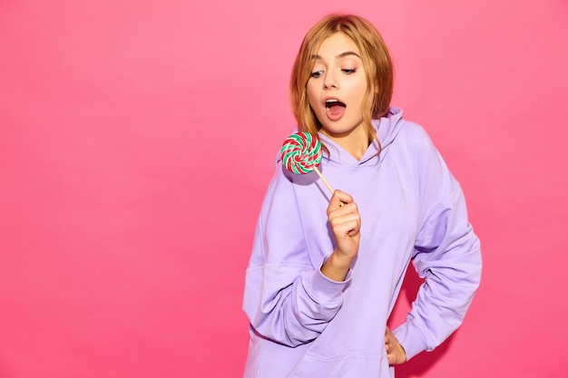 Portret van jonge mooie glimlachende hipster vrouw in trendy zomer hoodie. Het sexy onbezorgde vrouw stellen dichtbij roze muur. Positief model met lolly