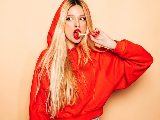 Portret van jonge mooie hipster slecht meisje in trendy rode hoodie en oorbel in haar neus. Positief model dat rond suikersuikergoed likt