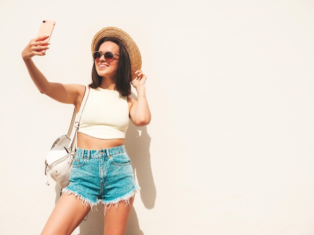 Portret van jonge mooie lachende hipster vrouw in trendy zomer jeans broek en hoed Sexy zorgeloos model poseren in de straat in de buurt van witte muur Positief model buitenshuis met werkmap Selfie nemen