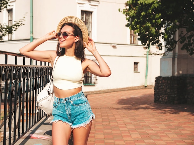 Gratis foto portret van jonge mooie lachende hipster vrouw in trendy zomer jeans broek en hoed sexy zorgeloos model poseren op de straat achtergrond bij zonsondergang positief model buitenshuis met werkmap