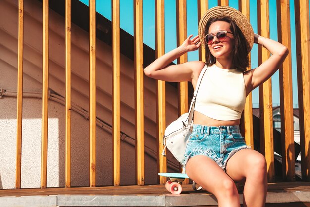 Portret van jonge mooie lachende hipster vrouw in trendy zomer jeans broek en hoed Sexy zorgeloos model poseren op de straat achtergrond bij zonsondergang positief model buitenshuis met werkmap