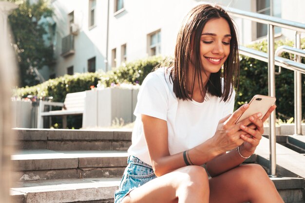 Portret van jonge mooie lachende hipster vrouw in trendy zomer jeans shorts Sexy zorgeloos model poseren in de straat zittend aan de trap kijken naar smartphone scherm met behulp van telefoon apps