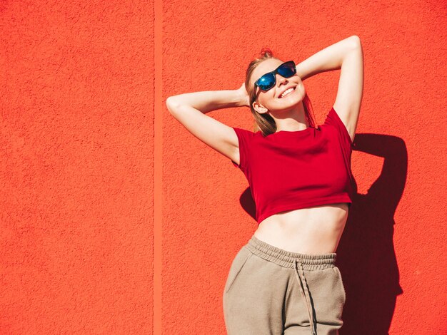 Portret van jonge mooie lachende hipster vrouw in trendy zomerkleren Sexy zorgeloze vrouw poseren in de buurt van rode muur in de straat bij zonsondergang Positief model buitenshuis Gelukkig en vrolijk