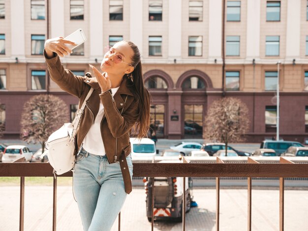 Portret van jonge mooie lachende meisje in zomer hipster jas en jeans. Model nemen selfie op smartphone. Vrouw maken van foto's in de straat. In zonnebril. Geeft luchtkus