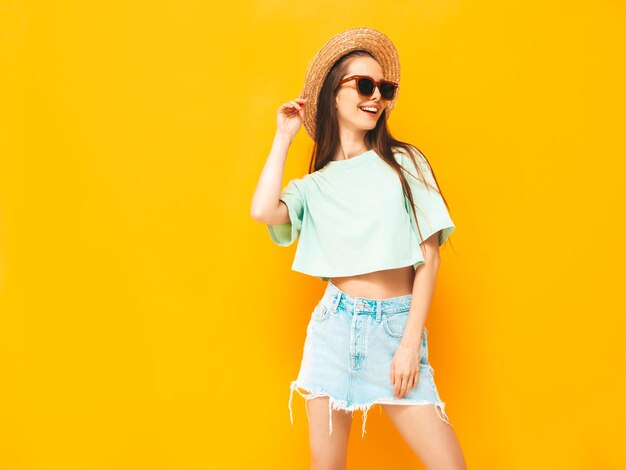 Portret van jonge mooie lachende vrouw in trendy zomer jeans rok zorgeloze vrouw poseren in de buurt van gele muur in studio Positief model plezier binnenshuis vrolijk en gelukkig In hoed