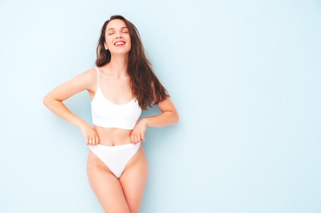 Portret van jonge mooie lachende vrouw in witte lingerie. sexy zorgeloos vrolijk model in ondergoed poseren in de buurt van lichtblauwe muur in studio. positieve en gelukkige vrouw die van de ochtend geniet