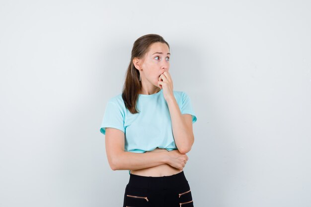 Portret van jonge mooie vrouw die haar nagels bijt, opzij kijkt in t-shirt, broek en gestrest vooraanzicht