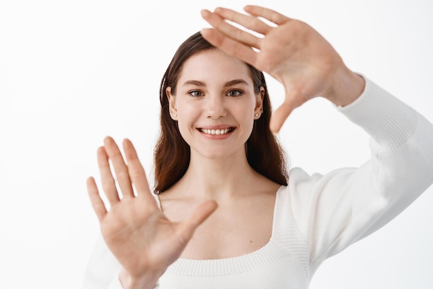 Portret van jonge mooie vrouw die vrolijk lacht en een cameraframe maakt met vingers Geïsoleerd op een witte achtergrond