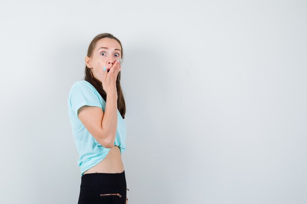 Portret van jonge mooie vrouw met hand op mond in t-shirt, broek en doodsbang vooraanzicht