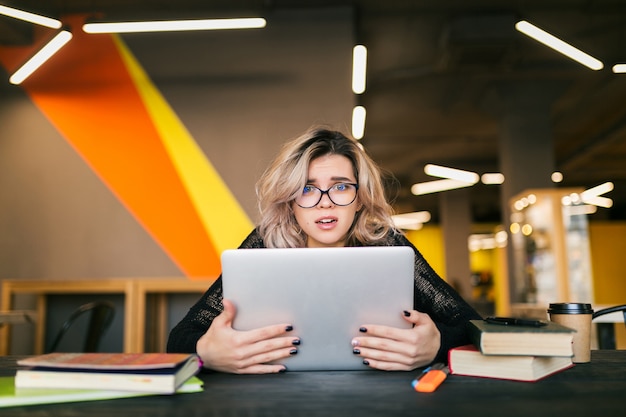 Portret van jonge mooie vrouw met socked gezichtsuitdrukking, zittend aan tafel bezig met laptop in co-working office, bril, stress op het werk, grappige emotie, student in de klas, frustratie