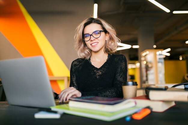 Portret van jonge mooie vrouwenzitting bij lijst in zwart overhemd die aan laptop in co-werkend bureau werken, die glazen dragen, glimlachend, gelukkig, positief