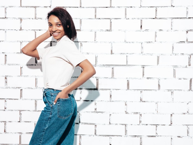 Portret van jonge mooie zwarte vrouw glimlachend model gekleed in zomer jeans en topkleren sexy zorgeloze vrouw poseren in de buurt van witte bakstenen muur in studio gelooid en vrolijk