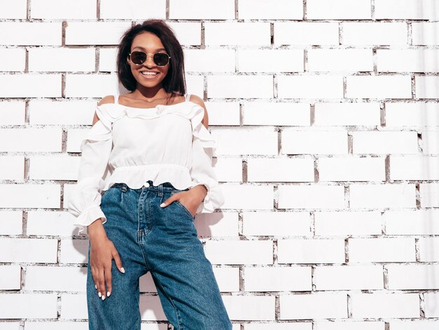 Portret van jonge mooie zwarte vrouw glimlachend model gekleed in zomer jeans kleding Sexy zorgeloze vrouw poseren in de buurt van witte bakstenen muur in studio gelooid en vrolijk