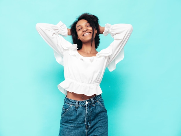 Portret van jonge mooie zwarte vrouw Glimlachend model gekleed in zomerse jeans kleding Sexy zorgeloze vrouw poseren in de buurt van blauwe muur in studio Gelooid en vrolijk