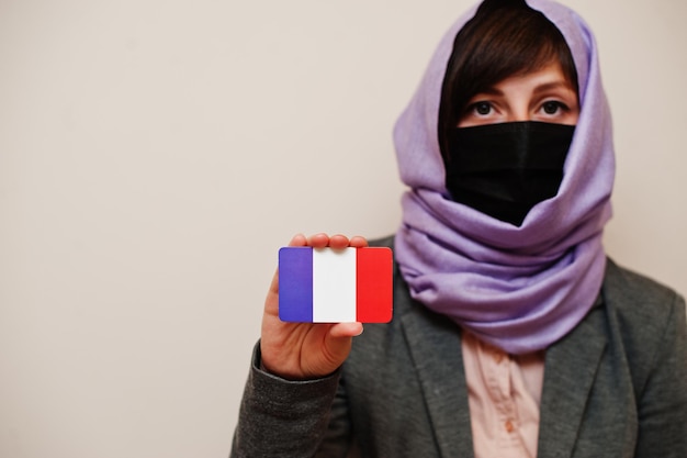 Gratis foto portret van jonge moslimvrouw die formele kleding draagt, beschermt gezichtsmasker en hijab-hoofddoek houdt de vlag van frankrijk vast tegen geïsoleerde achtergrond coronavirus landconcept