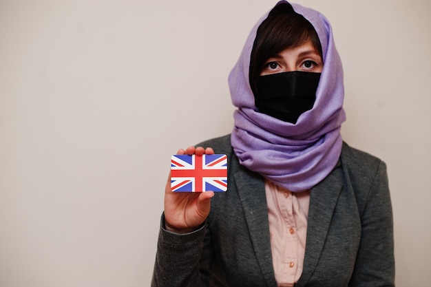 Portret van jonge moslimvrouw die formele kleding draagt, beschermt gezichtsmasker en hijab-hoofddoek houdt de vlag van het Verenigd Koninkrijk vast tegen geïsoleerde achtergrond Coronavirus landconcept