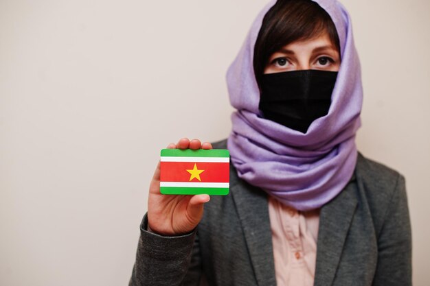 Portret van jonge moslimvrouw die formele kleding draagt, beschermt gezichtsmasker en hijab hoofddoek houdt Surinaamse vlagkaart tegen geïsoleerde achtergrond Coronavirus landconcept