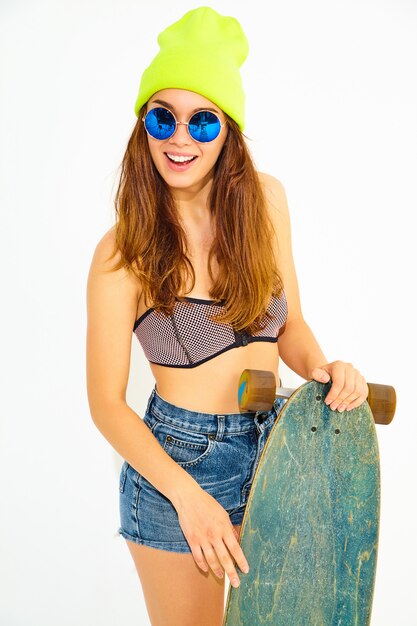 Portret van jonge stijlvolle lachende vrouw model in casual zomer badmode kleding en gele muts poseren met longboard bureau. Geïsoleerd op wit