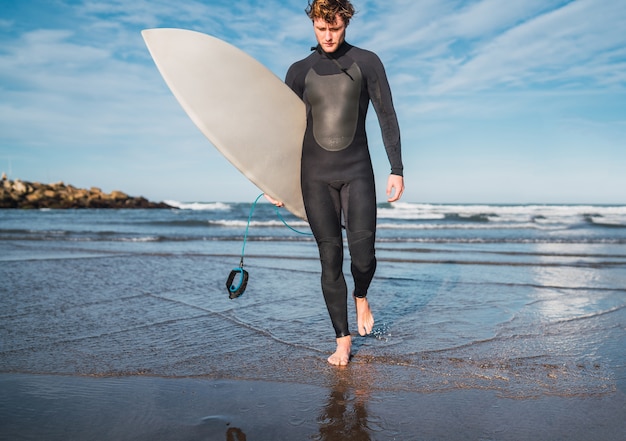 Portret van jonge surfer die het water verlaat met surfplank onder zijn arm. Sport en watersport concept.