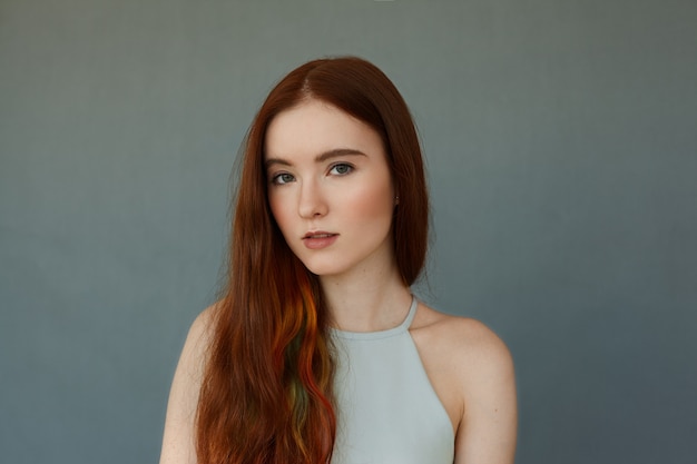Portret van jonge tedere roodharige tienermeisje met groene ogen dragen blauwe top kijken met ernstige of peinzende uitdrukking staande op blauwe muur, haar lippen lichtjes gescheiden