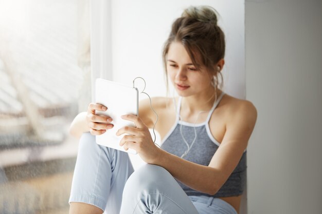 Portret van jonge tiener vrolijke vrouw in koptelefoon glimlachen kijken naar tablet surfen web surfen op internet zitten in de buurt van venster over witte muur.
