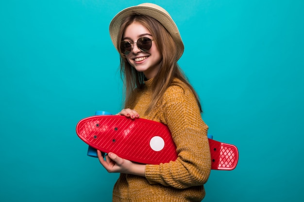 Portret van jonge tienervrouw in zonnebril en het skateboard van de hoedenholding terwijl status geïsoleerd over groene muur