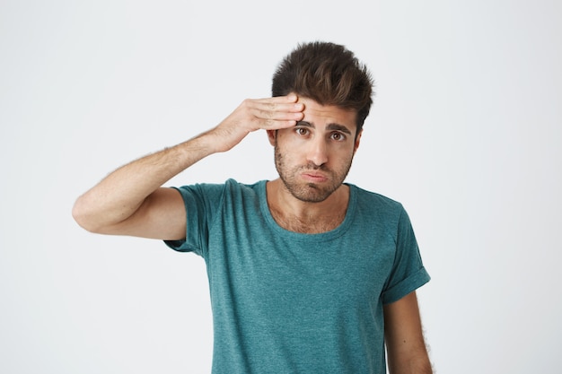 Portret van jonge vermoeide Kaukasische kerel die blauwe t-shirt draagt die zijn voorhoofd afveegt met hand die na complexe opleiding wordt vermoeid. Lichaamstaal.