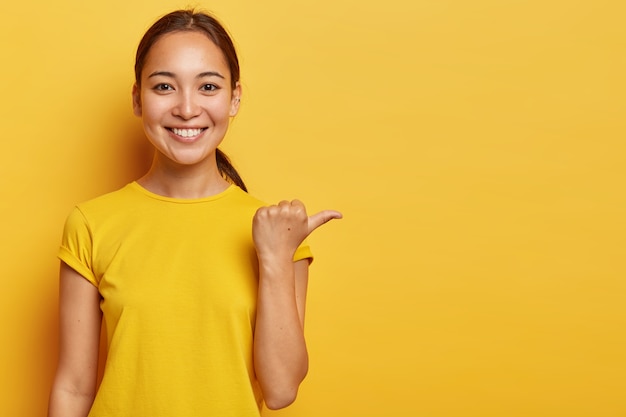 portret van jonge vrolijke Aziatische vrouw wijst weg met duim, blij gezichtsuitdrukking, toont kopie ruimte voor reclame, heeft een prettige uitstraling, draagt felgele kleding.