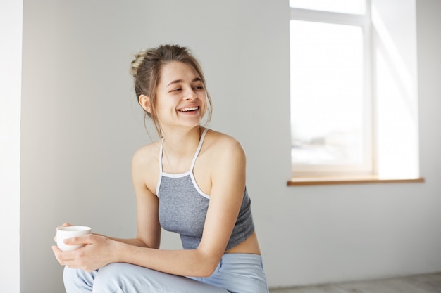 Portret van jonge vrolijke gelukkige vrouw die het lachen de zitting van de holdingskop op stoel over witte muur thuis glimlachen.