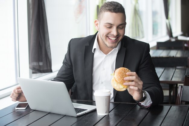 Portret van jonge vrolijke zakenman die hamburger eet terwijl hij in cafe werkt