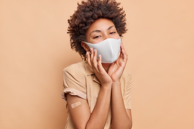 Portret van jonge vrouw met krullend haar houdt handen op het gezicht draagt beschermend masker pleister op arm wordt ingeënt tegen coronavirus geïsoleerd over beige muur