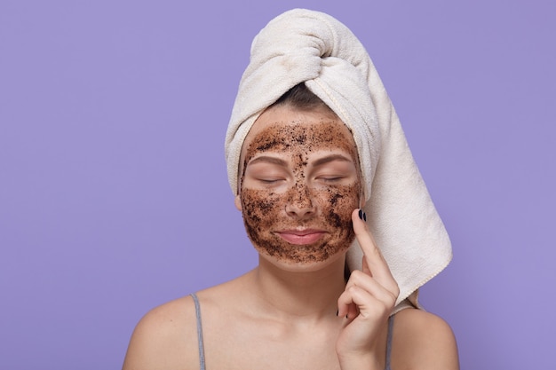 Portret van jonge vrouw past zelfgemaakte gezichts klei masker toe, heeft witte handdoek om het hoofd gewikkeld, de ogen gesloten